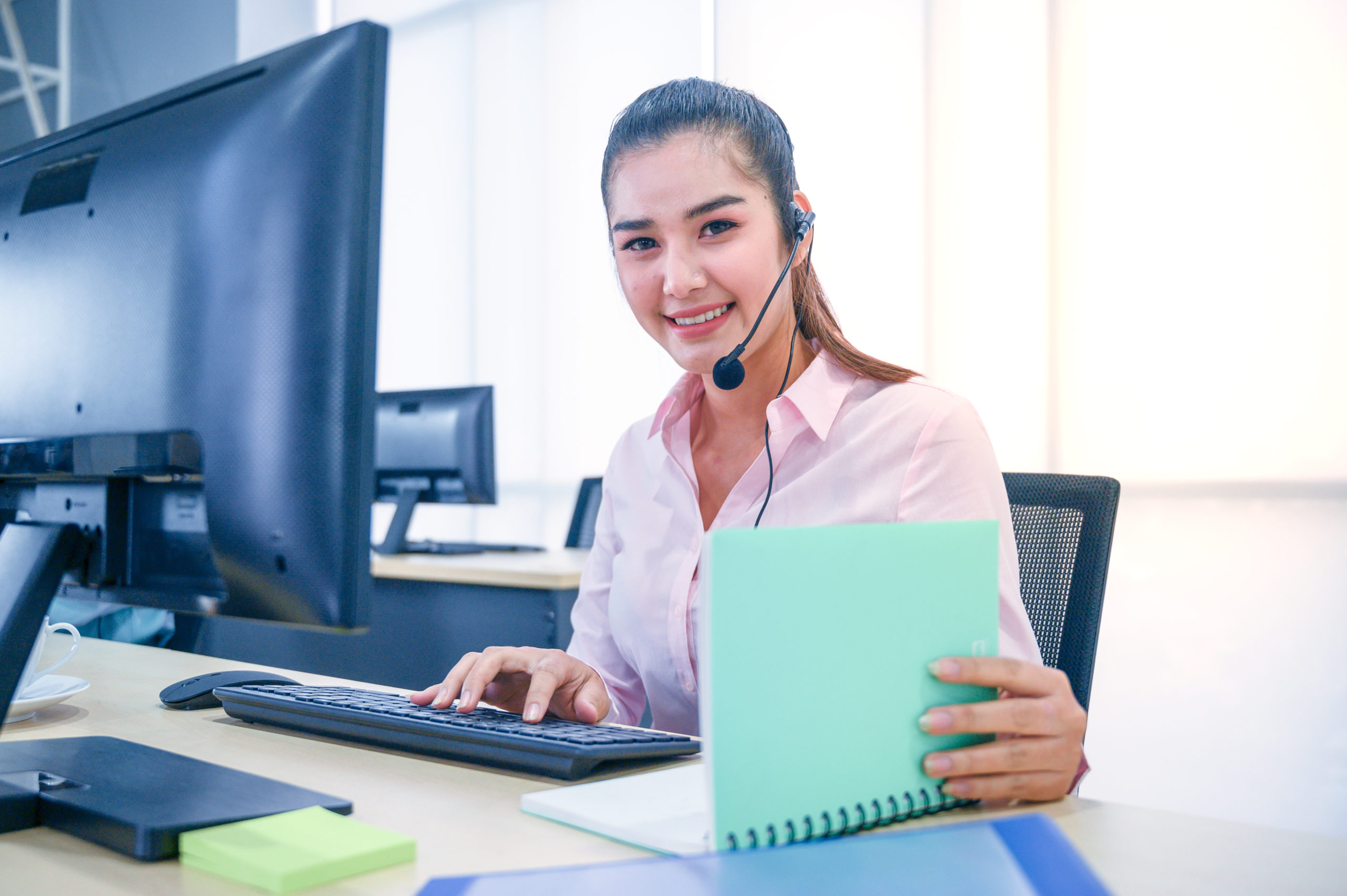 Smiling female customer service agents wearing a headset checks notes while working on a computer.