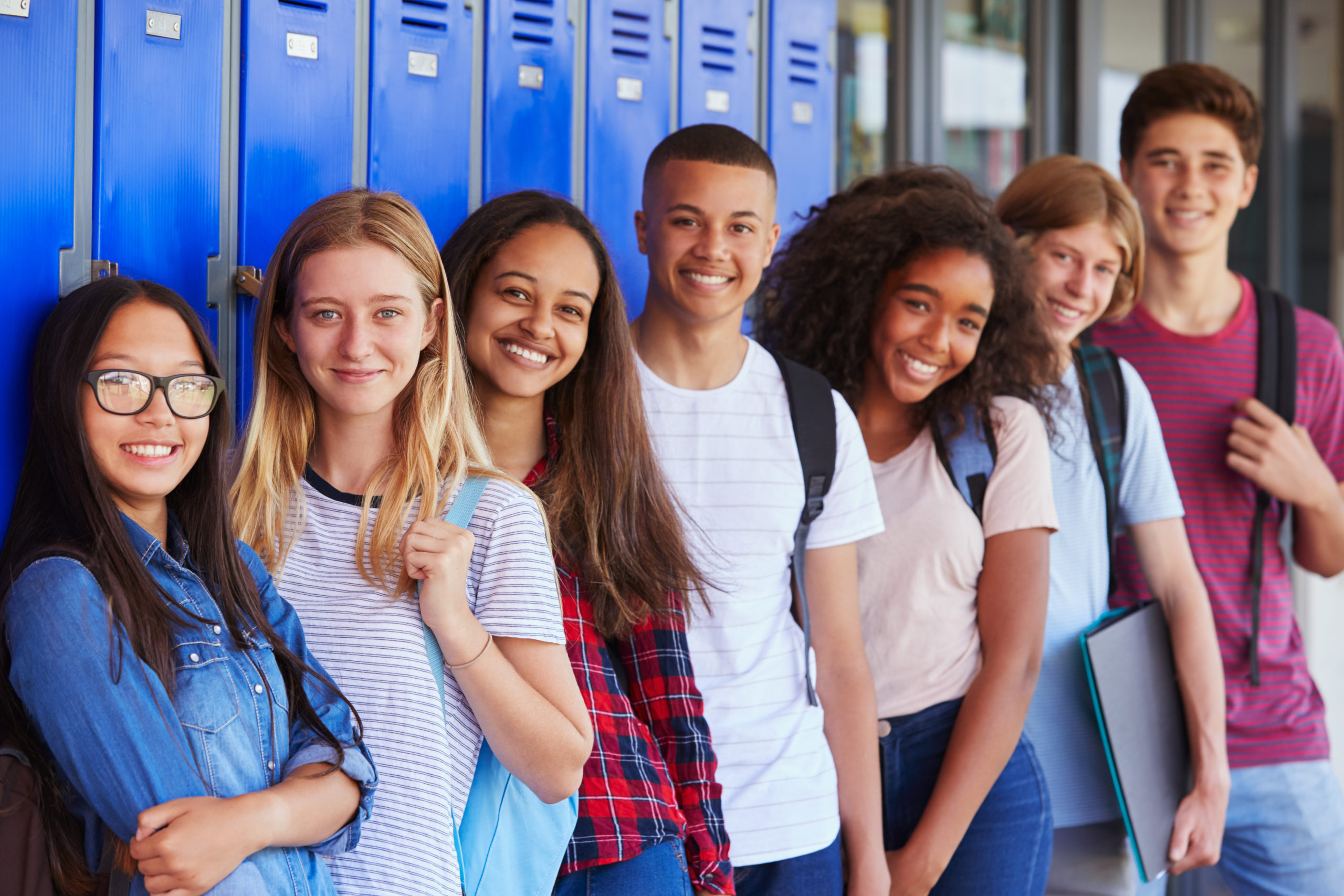 A diverse group of high school students leans against blue lockers in the hallway and smile. 