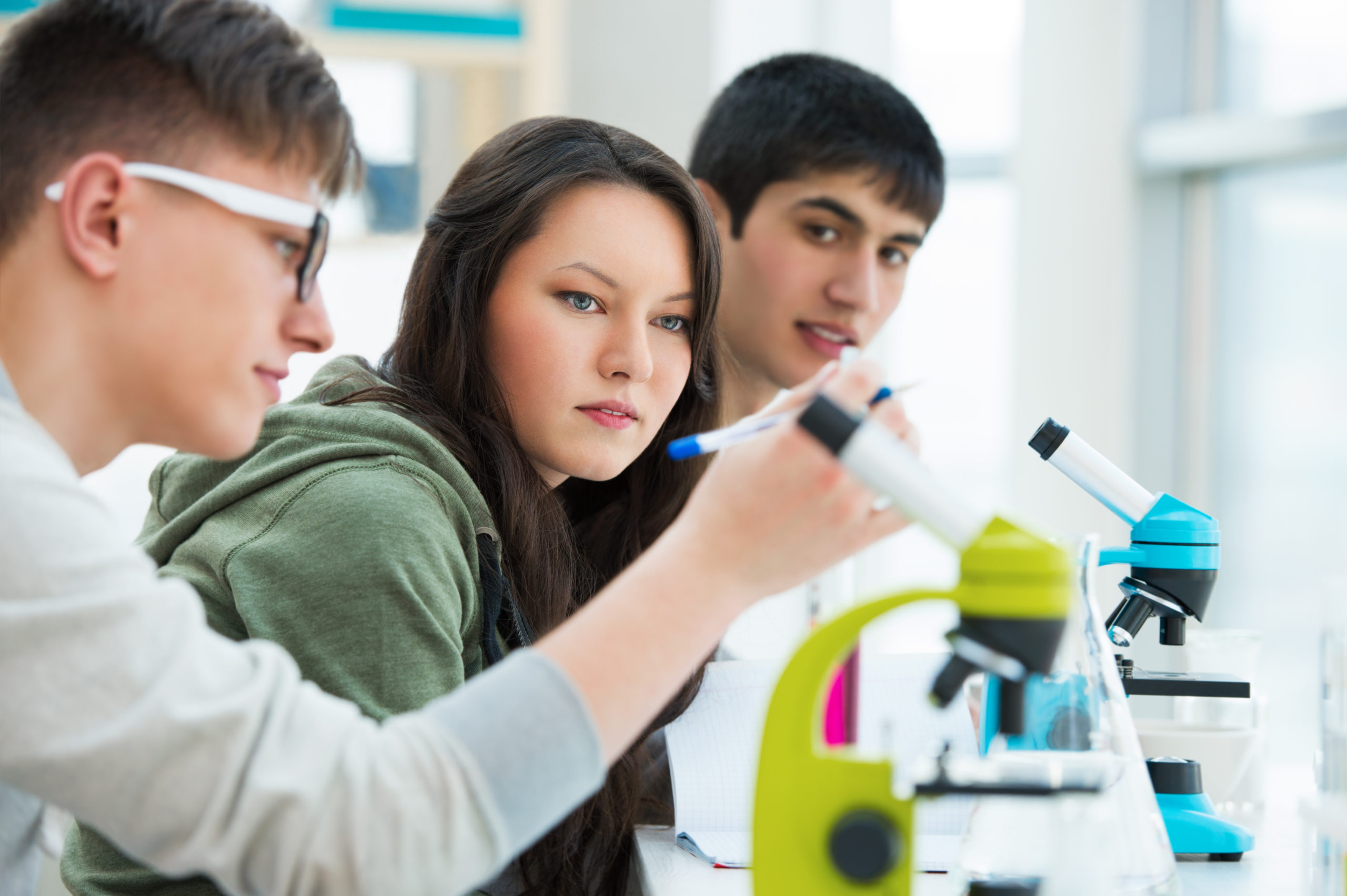  A girl and two boys in a high school science lab are preparing slides to look at through microscopes. 