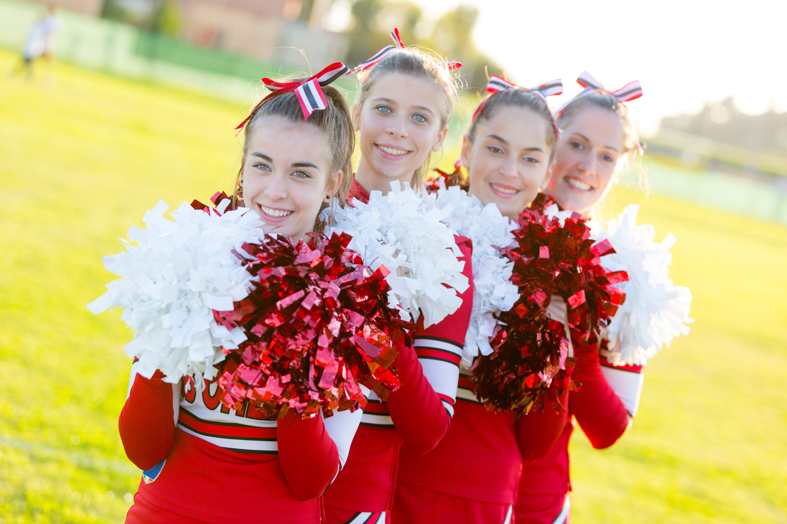  4 cheerleaders in red uniforms stand smiling in a line and holding pom-poms. 