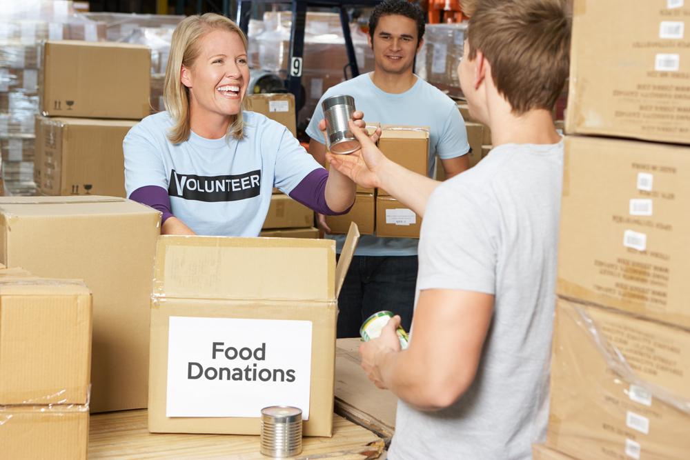 Young woman volunteer in a warehouse surrounded by food donation boxes takes cans from a young man. 