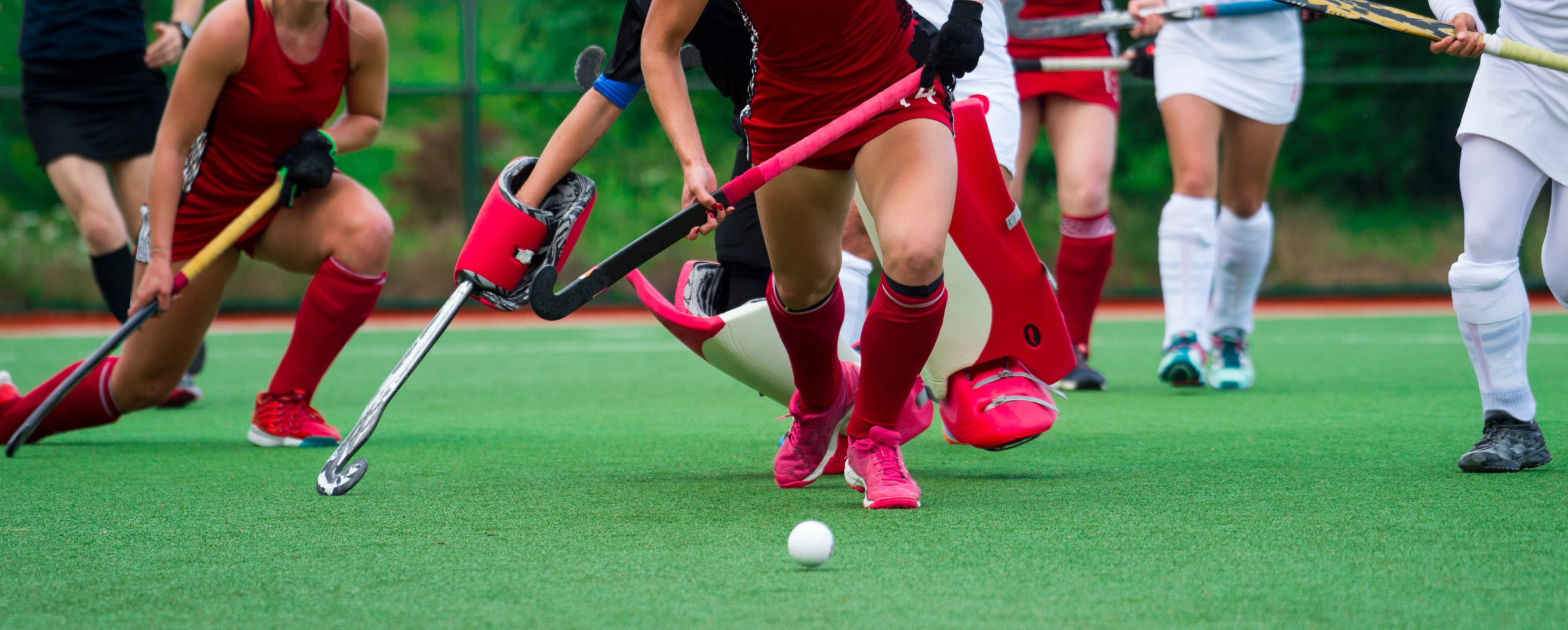Women’s field hockey players in white and red uniforms are playing and the red player has the ball. 
