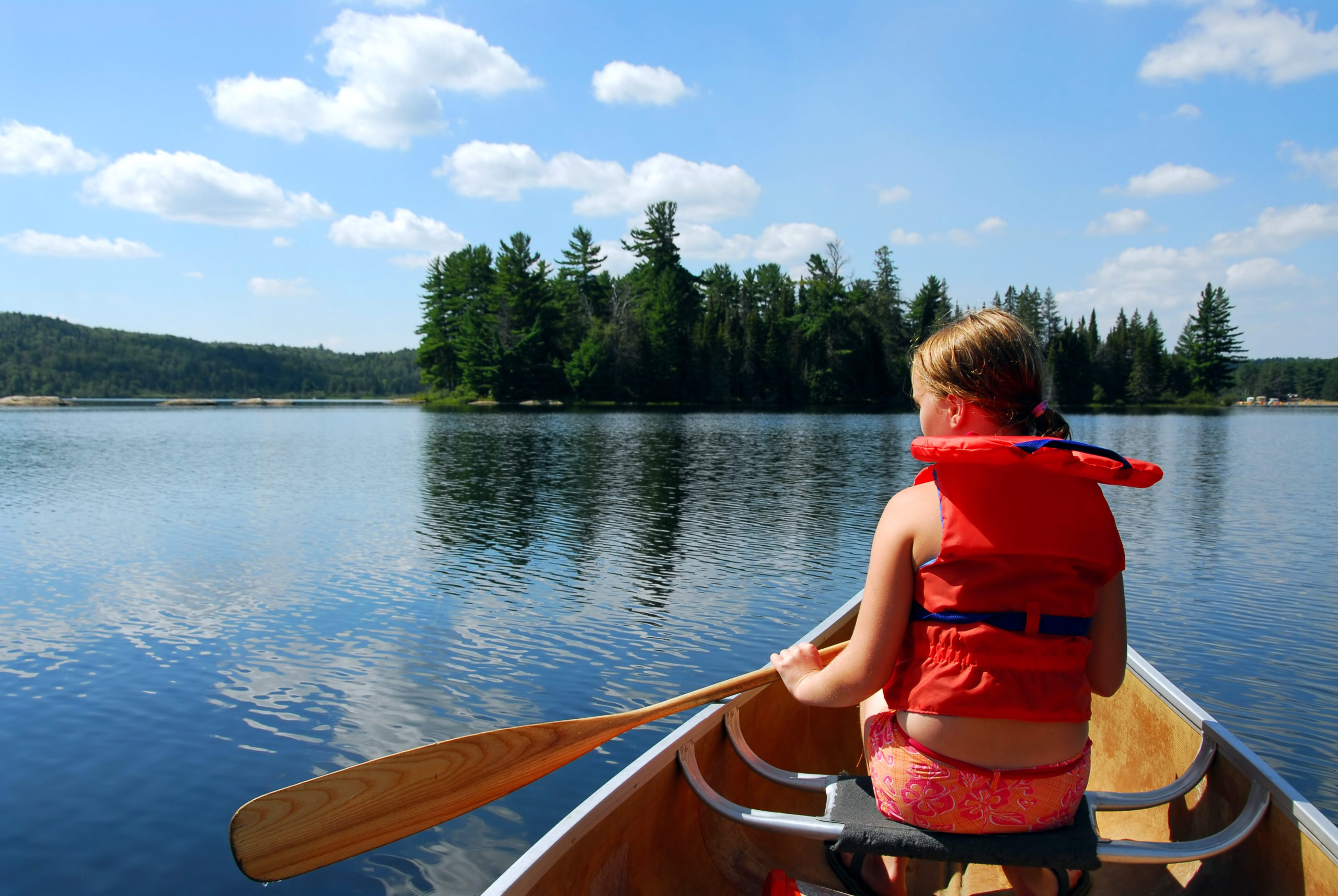 Young girl camper wearing a life vest is paddling in a canoe on a scenic lake. 