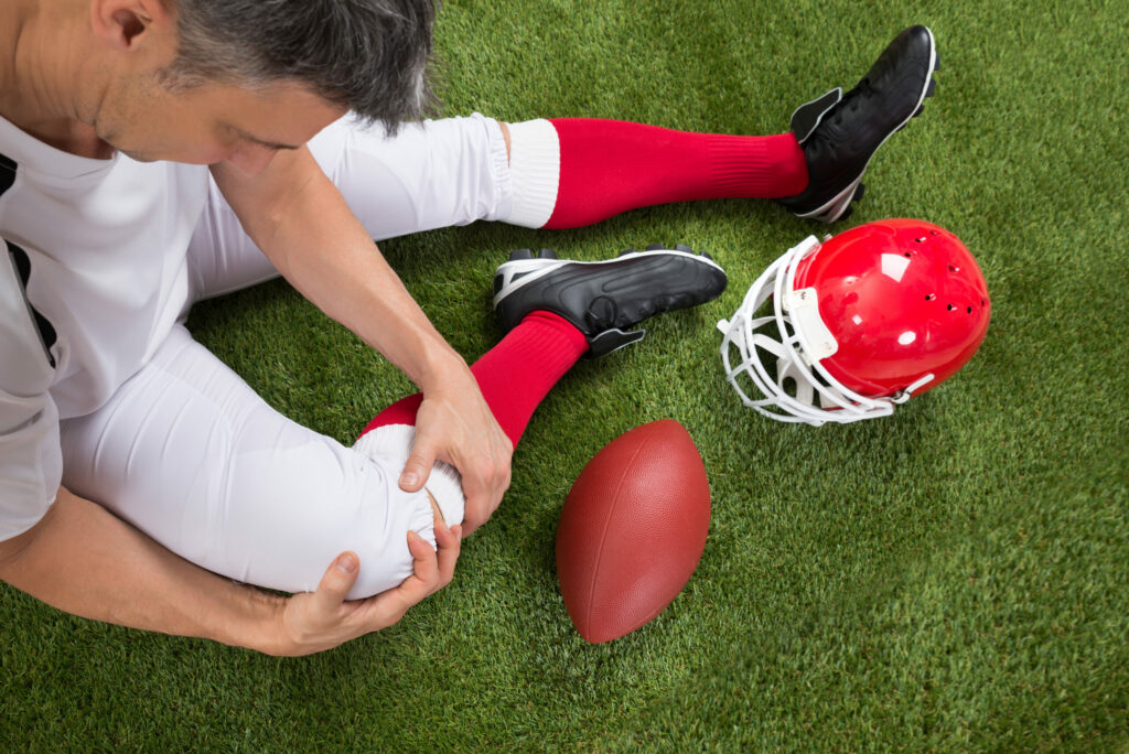 An injured football player sits on the ground next to his red helmet, suffering from a common sports knee injury