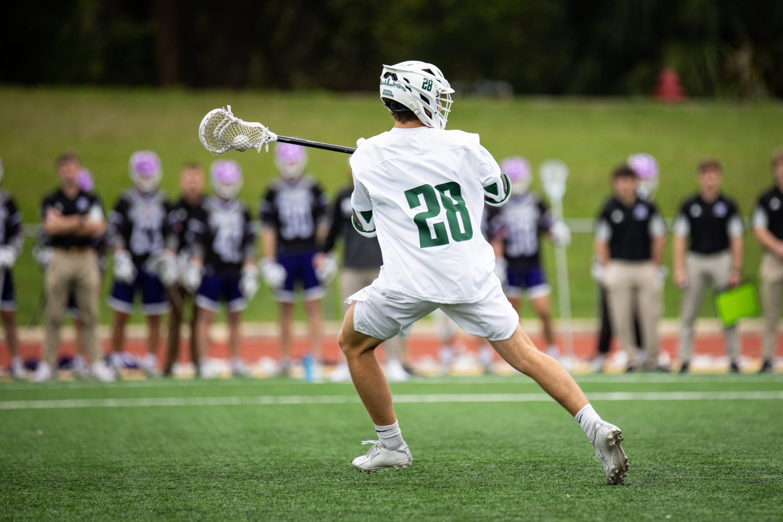 A college sports lacrosse player in white stands ready to pass the ball held in his stick’s pocket. 
