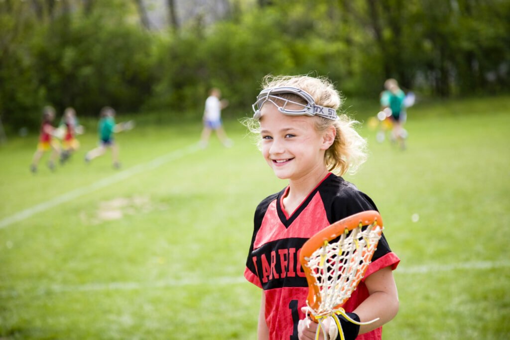 A young athlete covered by amateur sports insurance holds a lacrosse stick while the game plays in the background.
