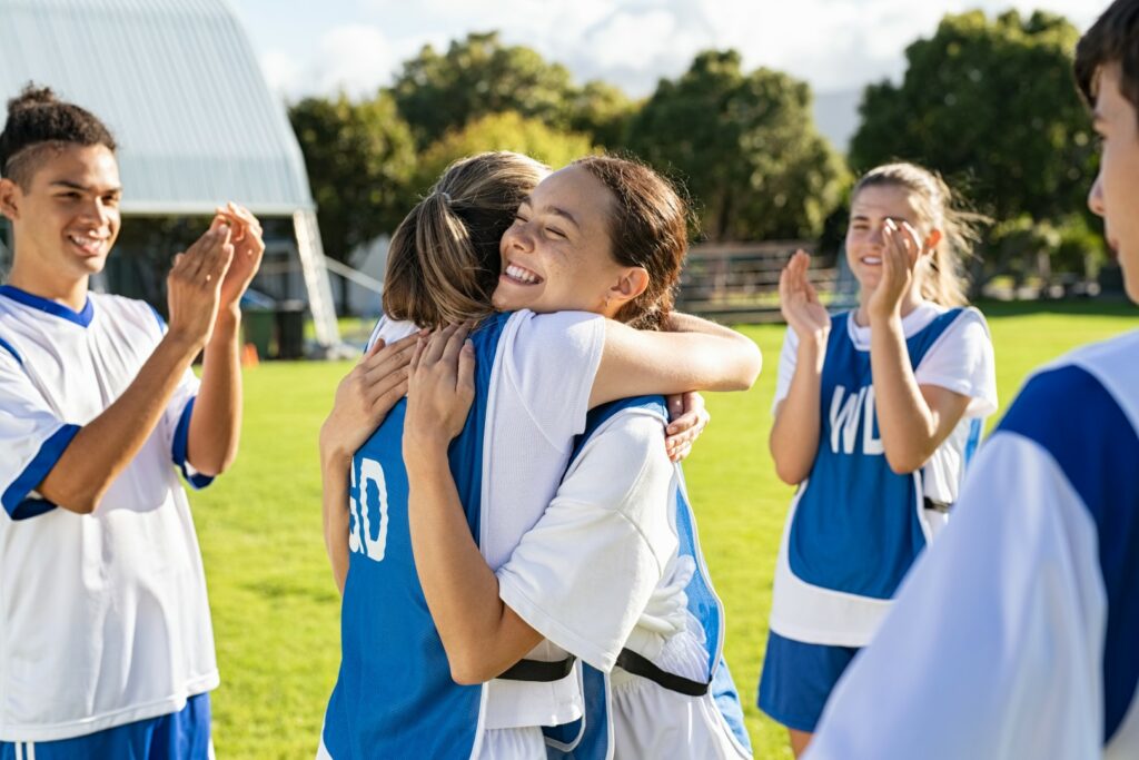 Happy high school soccer players embrace as teammates clap after their team wins.