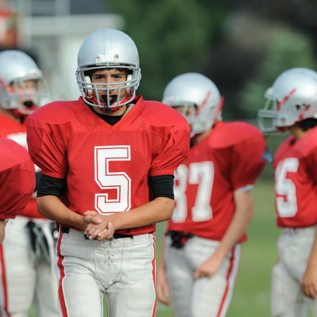 A young high school football player wearing a red jersey stands with his teammates on the field.